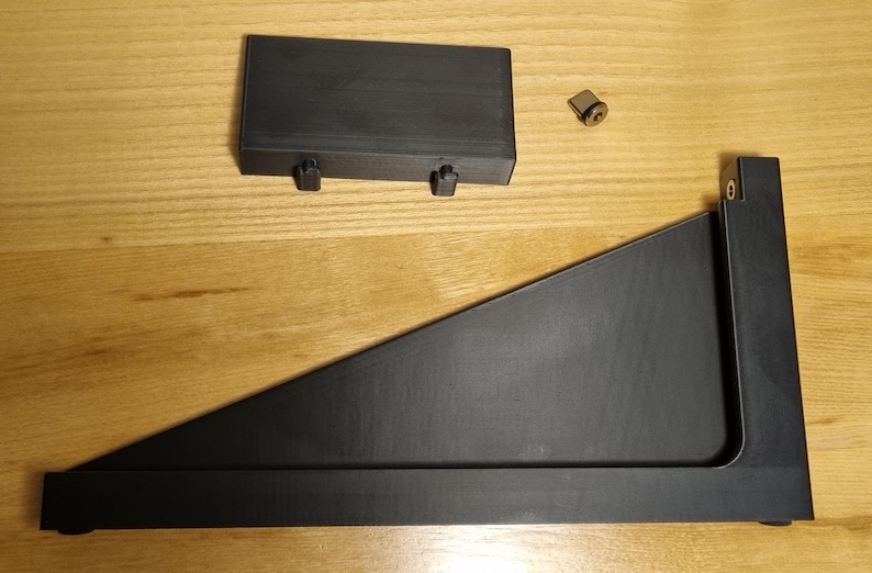 May include: Black wooden shelf bracket components, including a rectangular piece with two pegs, a triangular support, and a small metal fastener. The items are arranged on a light brown wooden surface.