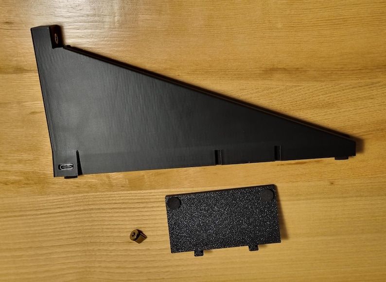 May include: Black, angled, electronic device stand with a rectangular base and a small, separate, textured black plate. A small, gold-colored screw is also present. The stand has a USB port.