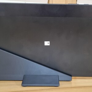 May include: A black tablet is propped up on a dark gray stand, with a small camera lens visible on the upper right corner. A white charging cable is plugged into the stand. The tablet is resting on a wooden surface.