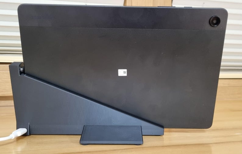 May include: A black tablet is propped up on a dark gray stand, with a small camera lens visible on the upper right corner. A white charging cable is plugged into the stand. The tablet is resting on a wooden surface.