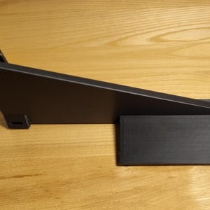 May include: A black, angled foot pedal with a rectangular base. The pedal has a hinged mechanism and is made of a matte black material. The item is set against a light brown wooden surface.