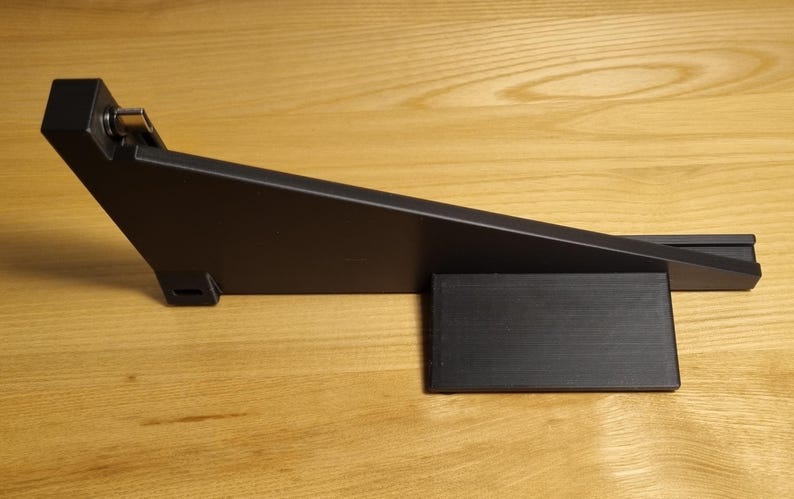 May include: A black, angled foot pedal with a rectangular base. The pedal has a hinged mechanism and is made of a matte black material. The item is set against a light brown wooden surface.