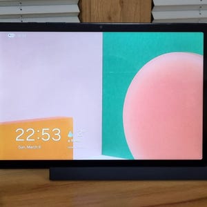 May include: A black tablet with a colourful display, resting on a black stand. The screen shows a pink circle, a green rectangle, and a yellow rectangle with the time 22:53 and the date Sunday, March 8th. The tablet is on a wooden surface.
