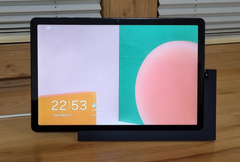 May include: A black tablet with a colorful display, showing a pink circle, a green rectangle, and a yellow rectangle with the time 22:53 and the date. The tablet is in a black stand on a wooden surface.