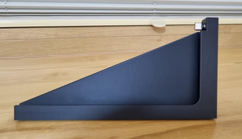May include: A black, angled shelf with a rectangular base and a triangular top. The shelf is made of a matte material and has a small, square metal accent on the right side. The shelf is designed to be mounted on a wall.