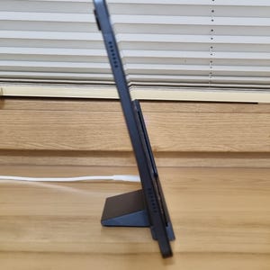 May include: A black tablet propped up on a matching stand, angled for viewing. The tablet is connected to a white charging cable. The background features a wooden surface and a window with horizontal blinds.