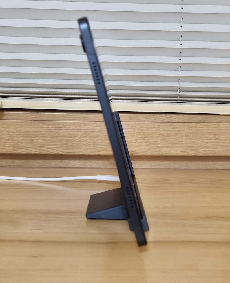 May include: A black tablet propped up on a matching stand, angled for viewing. The tablet is connected to a white charging cable. The background features a wooden surface and a window with horizontal blinds.