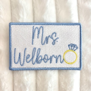Peut inclure: Écusson brodé avec le texte "Mrs. Welborn" en écriture bleu clair. Une bague jaune avec un diamant est à côté du nom. L'écusson a un fond blanc avec une bordure bleu clair.