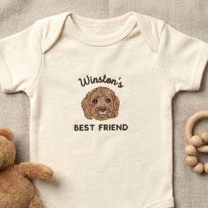 Op de afbeelding: Crèmekleurige babybody met de tekst "Winston's BEST FRIEND" en een bruine hondenillustratie. Een kleine bruine teddybeer en een houten bijtring liggen naast de body op een neutrale ondergrond.