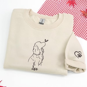 May include: A beige sweatshirt featuring a black line drawing of a dog with a butterfly. The dog's name, "Leo," is written below. The sleeve cuff has a heart and paw print design.