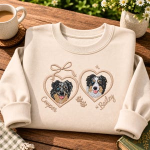 Può includere: Una felpa beige a girocollo con ricami di due cani pastore australiano all'interno di cornici a forma di cuore. Il cuore a sinistra ha un fiocco e il nome Cooper, mentre quello a destra presenta il nome Bailey e delle impronte di zampe.