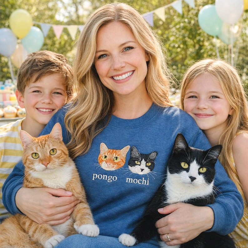 The item features multi-color embroidery on an indigo blue sweatshirt, with two custom cat portraits and their names, “pongo” and “mochi,” stitched on the middle chest for a personalized, realistic look.