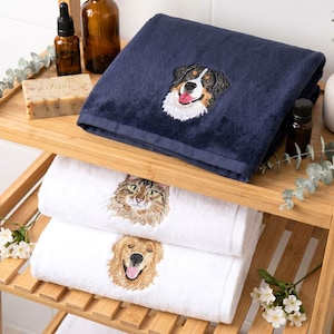 May include: A navy blue towel featuring an embroidered Bernese Mountain Dog head, stacked above two white towels, one with an embroidered brown tabby cat head and the other with an embroidered golden retriever head, all with detailed fur textures.
