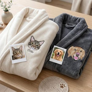 May include: Two plush bathrobes, one cream and one gray, are displayed. The cream robe features an embroidered cat face and a photo of a cat. The gray robe has an embroidered dog face and a photo of a dog. A coaster with a paw print is also visible.
