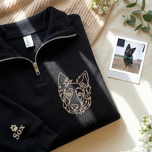 May include: Black quarter-zip sweatshirt with a gold-colored embroidered dog face design on the front. The left sleeve has a paw print and the name "Sox" embroidered in gold. A small photo of a dog is on the right.