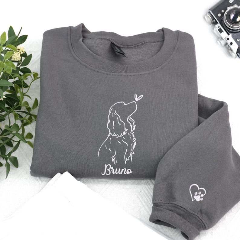 This charcoal sweatshirt shows a white outline of a Cocker Spaniel with a butterfly accent. The name “Bruno” is embroidered beneath, and a heart-shaped paw print decorates the sleeve.