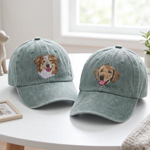 May include: Two sage green baseball caps with embroidered dog portraits. One cap features a brown and white dog, the other a golden retriever. Both caps have a curved brim and a top button.