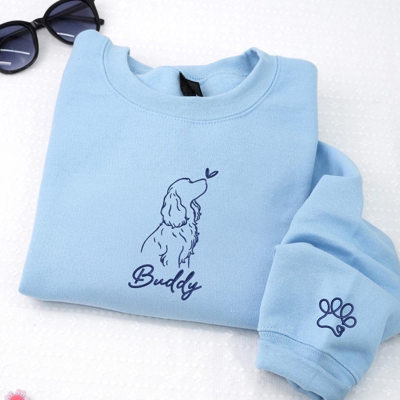 This light blue sweatshirt features a navy blue outline of a Cocker Spaniel with a butterfly above its nose. The name “Buddy” and a paw print on the sleeve complete the custom design.