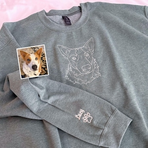 以下が含まれることがあります： 前面に犬の顔の白い輪郭が描かれたグレーのスウェットシャツ。袖には「Lucy」という名前と白い足跡が刺繍されています。犬の小さな写真が左上にあります。