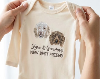Puede incluir: Body de bebé color crema con mangas largas. Presenta imágenes bordadas de dos perros y el texto "Zara & Gemma's NEW BEST FRIEND" en marrón oscuro. El body es sostenido por dos manos.
