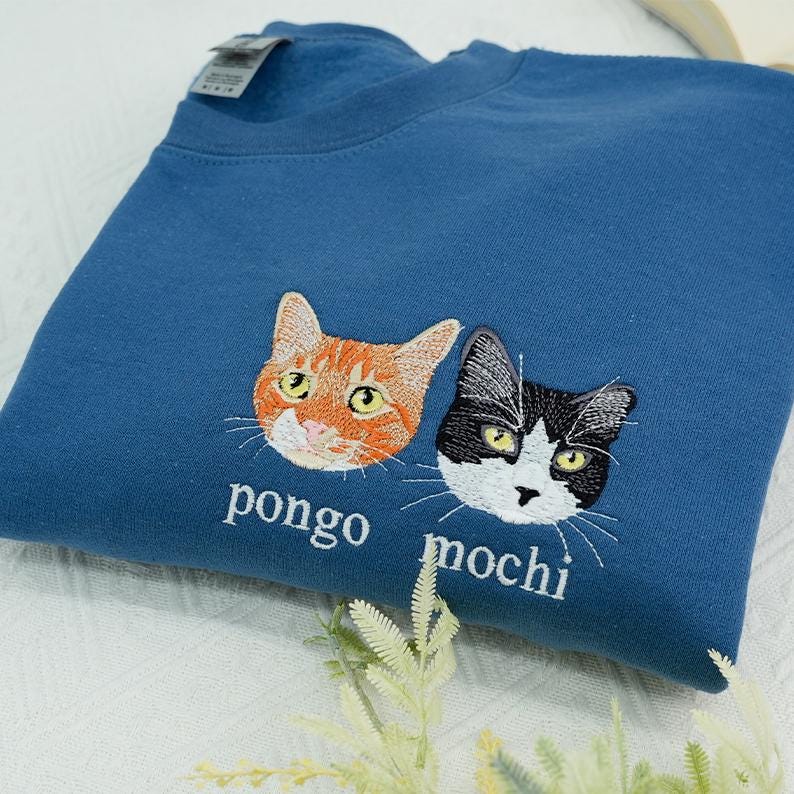 The item features multi-color embroidery on an indigo blue sweatshirt, with two custom cat portraits and their names, “pongo” and “mochi,” stitched on the middle chest for a personalized, realistic look.