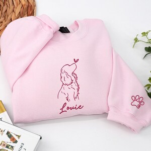 A light pink sweatshirt decorated with a maroon outline of a Cocker Spaniel gazing at a heart-shaped butterfly. The name “Louie” and a small paw print motif on the sleeve enhance the sweet detail