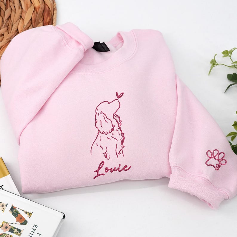 A light pink sweatshirt decorated with a maroon outline of a Cocker Spaniel gazing at a heart-shaped butterfly. The name “Louie” and a small paw print motif on the sleeve enhance the sweet detail