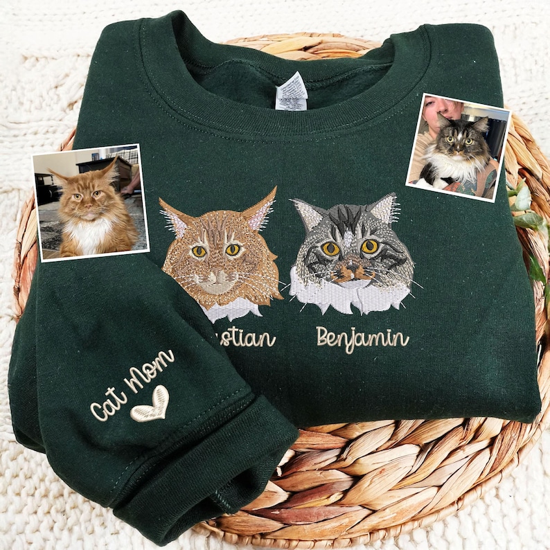 Forest green sweatshirt embroidered on the chest with two realistic cat portraits — a Maine Coon named Adrian and a Tabby Cat named Benjamin, stitched in detailed thread colors. On the right sleeve, the phrase Cat Mom is embroidered in beige with a small heart detail.