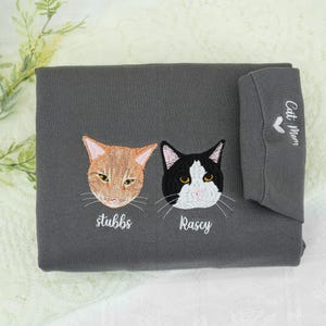 This charcoal gray sweatshirt features embroidered portraits of two adorable cats: Stubbs (orange) and Rascy (black & white), with their names stitched below. The sleeve adds a sweet touch with “Cat Mom” and a heart design — perfect for any devoted feline parent.