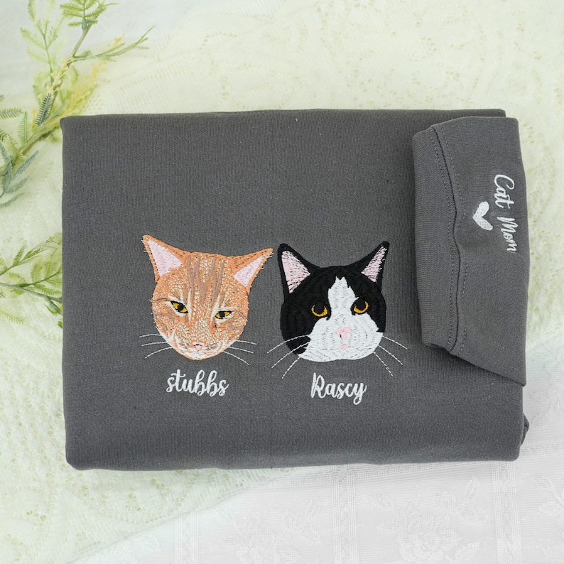 This charcoal gray sweatshirt features embroidered portraits of two adorable cats: Stubbs (orange) and Rascy (black & white), with their names stitched below. The sleeve adds a sweet touch with “Cat Mom” and a heart design — perfect for any devoted feline parent.