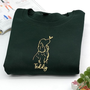 A forest green sweatshirt embroidered with a golden outline of a Cocker Spaniel looking up at a butterfly. The name “Teddy” is stitched below the design, adding a personalized touch.
