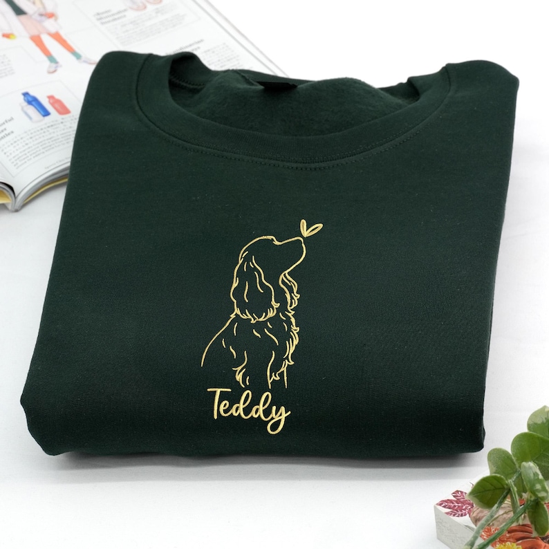 A forest green sweatshirt embroidered with a golden outline of a Cocker Spaniel looking up at a butterfly. The name “Teddy” is stitched below the design, adding a personalized touch.