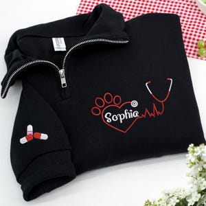Puede incluir: Sudadera negra con cremallera de un cuarto y cremallera blanca. La sudadera presenta un corazón bordado con huella de pata roja con el nombre "Sophia" y un diseño de estetoscopio. Un diseño de pastilla está bordado en la manga.