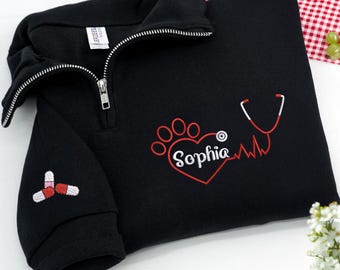 May include: Black quarter-zip sweatshirt with a white zipper. The sweatshirt features an embroidered red paw print heart with the name "Sophia" and a stethoscope design. A pill design is embroidered on the sleeve.