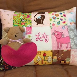 May include: A pink and brown patchwork pillow with a brown teddy bear sleeping on a pink heart. The pillow has the name "Lily" embroidered on it.