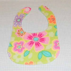 May include: A green baby bib with a floral pattern featuring pink, yellow, orange, and purple flowers. The bib has a white trim around the edges.