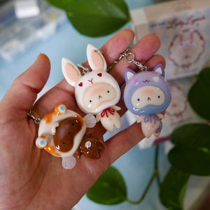 May include: Three small, glossy, character-themed keychains featuring a brown and white patterned creature, a white rabbit-hooded figure with red heart accents, and a lavender cat-hooded figure, each attached to a silver metal jump ring.