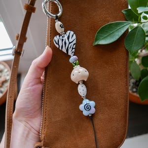 Könnte beinhalten: Ein dekorativer Taschenanhänger mit einem silberfarbenen Ring, einer cremefarbenen Perle mit kleiner Blume, einer herzförmigen Perle mit schwarz-weißem Zebramuster, einer hellblauen Perle, einer beigen, kreaturförmigen Perle, einer weißen Perle mit schwarzen Punkten und einer hellblauen, blumenförmigen Perle mit schwarzer Spirale in der Mitte.