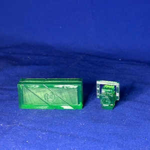 Puede incluir: Dos objetos de plástico verde impresos en 3D. Uno es un bloque rectangular con un diseño en relieve y el otro es una pequeña figura detallada.