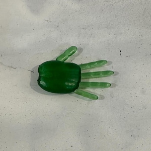 Puede incluir: Un objeto verde en forma de mano con cinco dedos. La palma es de un tono verde más oscuro que los dedos. El objeto probablemente está hecho de un material translúcido, posiblemente vidrio o resina.