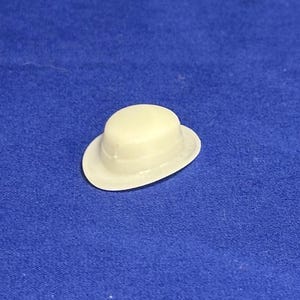 May include: A small, white, plastic hat with a rounded crown and a brim. The hat is a miniature version of a traditional bowler hat.