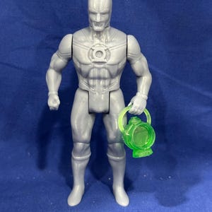 Op de afbeelding: Een grijs actiefiguur van een superheld, met een groene lantaarn. Het figuur staat in een pose, met een gedetailleerd kostuum en een serieuze uitdrukking. De groene lantaarn is doorschijnend.