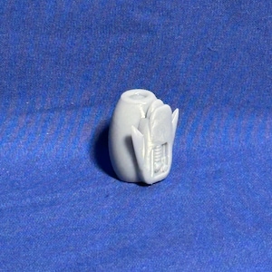 May include: A light gray, egg-shaped object with a detailed, textured design on one side. The object has a cylindrical opening at the top and appears to be a decorative item or a small sculpture. The background is a solid blue fabric.