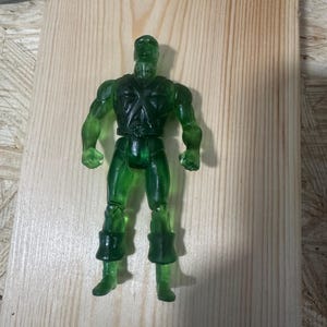 Puede incluir: Una figura de acción verde translúcida de un personaje humanoide musculoso. La figura tiene un diseño detallado, que incluye un emblema en forma de cruz en el pecho y botas segmentadas. Se encuentra sobre una superficie de madera clara.