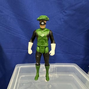 Puede incluir: Una figura de acción de Green Lantern con una cabeza de brócoli. La figura lleva un traje verde y negro con guantes blancos y botas verdes. El símbolo de Green Lantern está en el pecho. La figura está en un recipiente de plástico transparente.