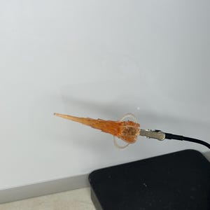 Peut inclure: Un petit objet orange en forme de flamme avec un anneau transparent et une pince métallique attachée à un fil noir. L'objet est posé sur un fond blanc et un carré noir.