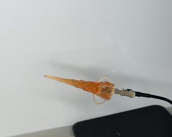 May include: A small, orange, flame-shaped object with a clear ring and a metal clip attached to a black wire. The object is set against a white background and a black square.