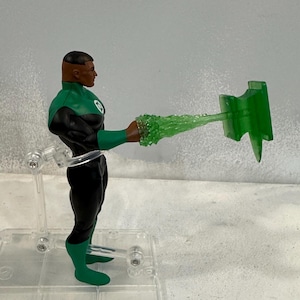 May include: A green and black action figure of a superhero holding a green energy construct. The figure is posed with its arm extended, and the energy construct is shaped like a beam. The figure is standing on a clear plastic stand.