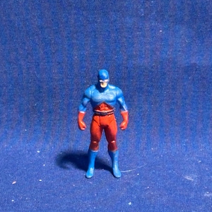 Pode incluir: Uma pequena figura de a&ccedil;&atilde;o de super-her&oacute;i com um esquema de cores azul e vermelho. A figura tem uma m&aacute;scara, peito e botas azuis, com bra&ccedil;os e pernas vermelhas. A figura est&aacute; sobre uma superf&iacute;cie azul.
