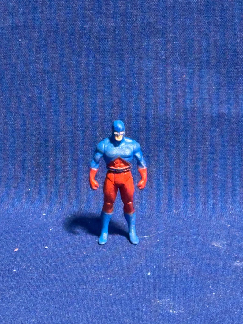 Pode incluir: Uma pequena figura de a&ccedil;&atilde;o de super-her&oacute;i com um esquema de cores azul e vermelho. A figura tem uma m&aacute;scara, peito e botas azuis, com bra&ccedil;os e pernas vermelhas. A figura est&aacute; sobre uma superf&iacute;cie azul.